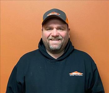 Photo of a man in a black hat standing against an orange background.  He has a black sweatshirt on with the SERVPRO logo